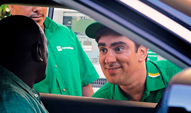 Imagem de dentro de um carro, com Marcelo Adnet vestido de frentista dos Postos Petrobras, do lado de fora da janela, abaixado, falando com o motorista