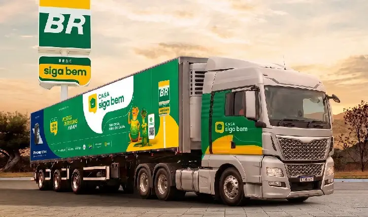Caminhão de carga da rede BR Mania estacionado em área externa ao entardecer. O veículo está adesivado com a identidade visual da campanha “Casa Siga Bem”, em tons de verde, amarelo e branco, com ilustrações e mensagens promocionais nas laterais. Ao fundo, aparece um totem dos Postos BR com a marca “Rede Siga Bem”.